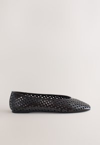 Chaussure plate noire avec motif perforé carré et bout arrondi, vue de profil sur un fond neutre.