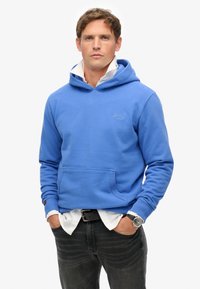 Felpa blu realizzata in tessuto morbido, con tasca frontale, cappuccio regolabile e polsini a coste. Indossata sopra una camicia bianca con colletto e jeans scuri.