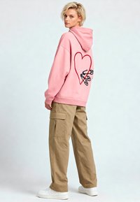 Felpa con cappuccio rosa con grande cuore rosso e contorno nero sul retro, abbinata a pantaloni cargo beige e sneakers bianche.