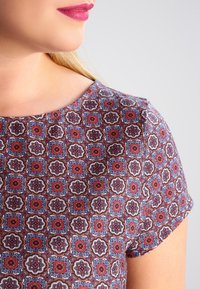 Top de manga corta en tejido estampado con diseños geométricos en rojo, azul y marrón. Textura suave con escote redondo.