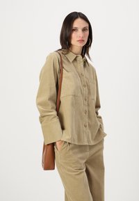 Beige kordfløyelskjorte med krage, knappelukking foran og to brystlommer. Kombinert med matchende bukser og holder en brun skinnveske.