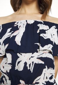 Femme portant un haut bleu marine à épaules dénudées avec de grandes impressions florales rose clair et blanche et un col à volants.