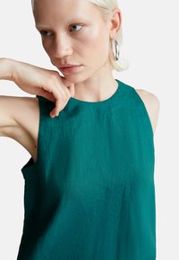 Tyrkysový bezrukávový top vyrobený z texturované látky. Má kulatý výstřih a minimalistický design, s viditelnými švy.