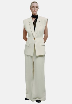 Femme portant un blazer sans manches crème oversize à un bouton, un pantalon large assorti, un top noir transparent et des chaussures noires.