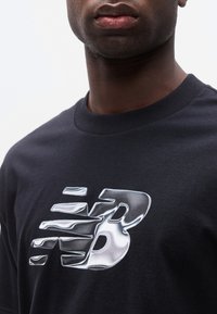 Κοντινό πλάνο ενός ατόμου που φοράει μαύρο πουκάμισο με ένα ανακλαστικό ασημί στυλιζαρισμένο γράμμα "B" στο στήθος, σε απλό φόντο.