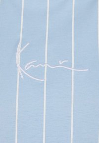 Lyseblå stripete stoff med vertikale hvite linjer. Har brodert signatur i hvitt. Glatt tekstur, uformell design.