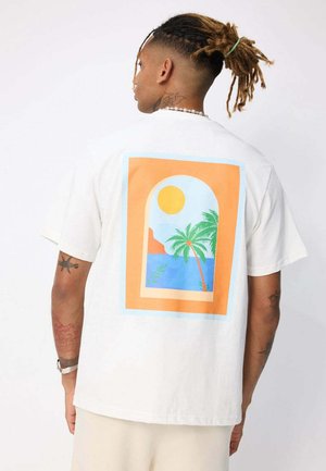 Homme avec des dreadlocks portant un t-shirt blanc affichant un motif coloré de palmier, soleil et océan vus à travers une fenêtre en arc dans le dos.