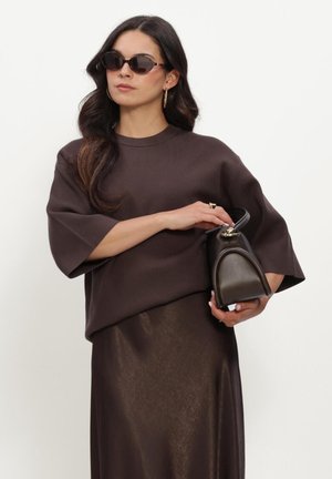 Woman with long dark hair wearing dark brown top and skirt, holding a small dark leather handbag, and wearing dark oval sunglasses.