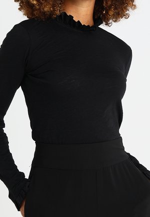 Kobieta ubrana w dopasowaną czarną bluzkę z długim rękawem, ozdobioną falbanką przy wysokim kołnierzu, oraz czarne, dopasowane spodnie z wysokim stanem.