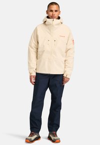 Beige veden-, mutta ei tuulenpitävä takki, jossa on kaksi edessä olevaa vetoketjullista taskua, säädettävä huppu ja erottuva oranssi logo. Yhdistettynä tummansinisiin työshortseihin ja harmaisiin kenkiin.