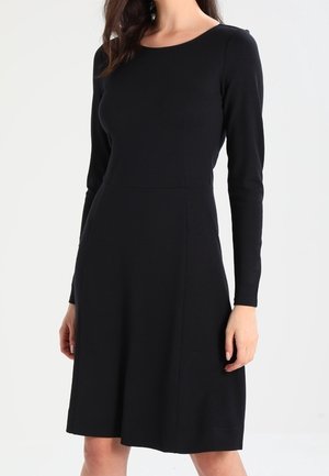 Woman wearing a plain black long-sleeve dress with a fitted waist and knee-length A-line skirt against a white background.