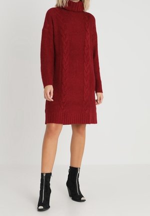 Femme portant une robe-pull à col roulé en maille torsadée rouge et des bottines noires à talons avec des fermetures éclair sur l'avant sur un fond clair.