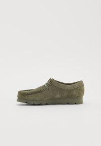 Chaussure en daim vert olive avec un bout rond, des coutures décoratives et une semelle bicolore. Présente un détail de lacet à l'avant et une silhouette basse.