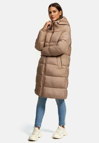 Long beige puffer coat with a hood, featuring horizontal quilted sections, zip closure, and an accent pocket. Worn over blue jeans and sneakers.