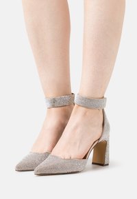 Chaussures à talons hauts argentées à bout pointu, ornées de strass scintillants, avec une bride épaisse à la cheville et un talon carré.