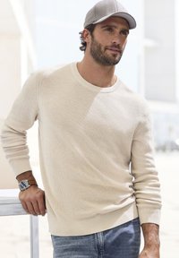 Beige strukturiertes Strickoberteil mit Rundhalsausschnitt, kombiniert mit blauer Denim. Eine silberne Armbanduhr ist am linken Handgelenk sichtbar.