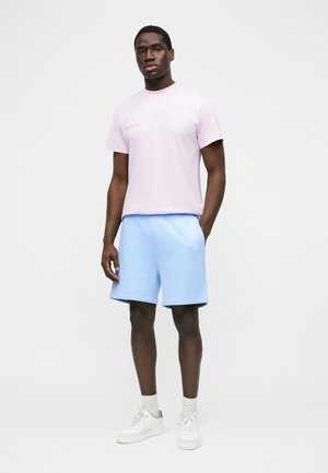 Man staat tegen een witte achtergrond en draagt een lichtpaars T-shirt, lichtblauwe korte broek, witte sokken en witte sneakers.