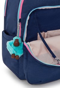 Mochila azul marina con cremalleras rosas y turquesas, que presenta un llavero de mono de peluche turquesa. El interior tiene un forro beige y múltiples bolsillos.