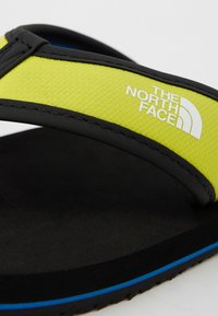 Juodos šlepetės su tekstūruotu ryškiai geltonu dirželiu, ant kurio yra balta "The North Face" logotipas, ir mėlynas akcentas palei pado kraštą.
