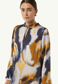 Langärmliges Hemd mit hohem Kragen, ausgestattet mit einem Schlüsselloch-Ausschnitt, in Tönen von Marineblau, Orange und Creme mit einem fließenden, abstrakten Muster.