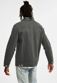 Grå denimjacka med krage, knäppta �ärmslut och sidofickor, med en strukturerad design och synliga sömmar på baksidan.