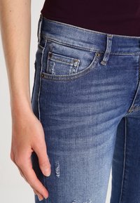 Jeans i denim med en urtvättad blå nyans, med slitna detaljer, femfickorsdesign och mässingsdetaljer. En hand vilar på midjebandet.