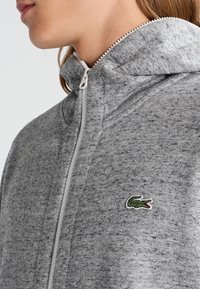 Sudadera con cremallera gris en tela mezcla, con un cuello alto y un pequeño logo de cocodrilo bordado en verde en el lado izquierdo.