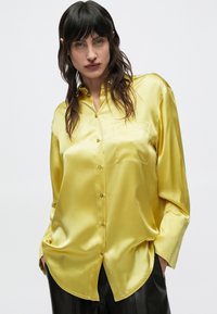 Femeie cu păr lung și întunecat, purtând o cămașă lejeră, lucioasă, galben satin cu nasturi și pantaloni negri, stând cu mâinile în buzunare.