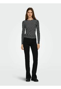 Schwarzes langärmliges Oberteil mit weißen Polka Dots, kombiniert mit schwarzen ausgestellten Hosen und klobigen schwarzen Schuhen. Einfacher runder Ausschnitt, taillierte Passform.