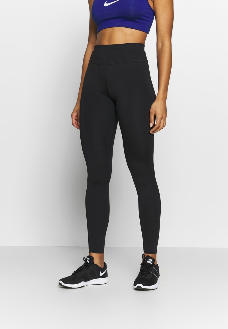 Leggings neri realizzati in un materiale elastico con una fascia alta in vita, abbinati a un crop top blu con un logo bianco. Scarpe da ginnastica nere completano l'outfit.