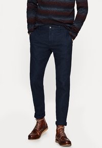 Granatowe chinosy wykonane z gładkiego materiału, posiadające klasyczny krój, kieszenie z przodu i zapięcie na guzik. Noszone z brązowymi skórzanymi butami.