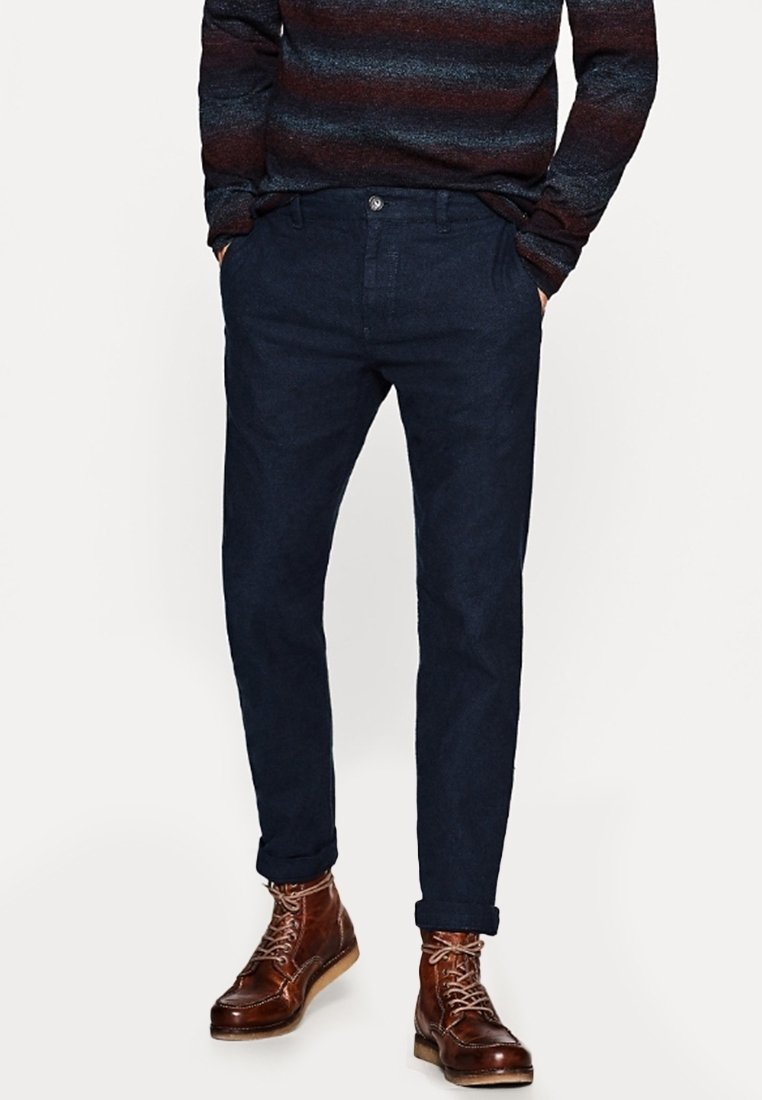 Granatowe chinosy wykonane z gładkiego materiału, posiadające klasyczny krój, kieszenie z przodu i zapięcie na guzik. Noszone z brązowymi skórzanymi butami.