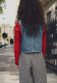 Chaleco de mezclilla sobre una camisa de manga larga roja combinado con pantalones de pierna ancha a cuadros en blanco y negro. Cabello rizado visible desde atrás.