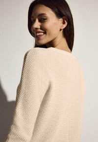 Waffelstruktur, hellbeige Pullover mit rundem Halsausschnitt und langen Ärmeln. Der Stoff sieht weich und leicht dehnbar aus.