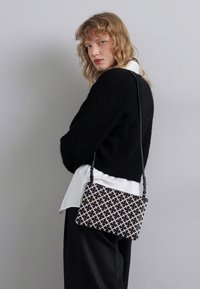 Maglione nero cropped sopra una camicia bianca, con una borsa geometrica nera e bianca a motivo croce e una sottile tracolla.