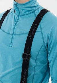 Nahaufnahme einer Person, die ein blaues, mit Reißverschluss versehenes Sportoberteil trägt, während sie schwarze Träger mit dem Whistler-Logo über die Schultern hat.