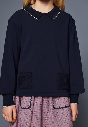 Navy long-sleeve top with white-trimmed collar and cuffs paired with pink checkered skirt featuring navy scalloped-trimmed pockets.