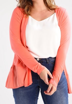 Femme portant un cardigan corail à manches longues par-dessus un top blanc et un jean bleu, les mains doucement jointes devant.