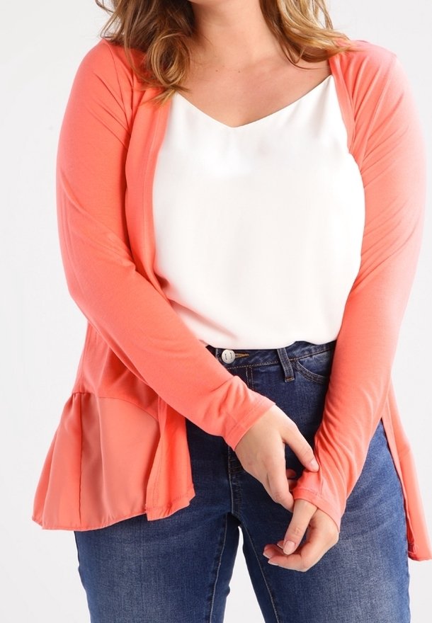 Femme portant un cardigan corail à manches longues par-dessus un top blanc et un jean bleu, les mains doucement jointes devant.