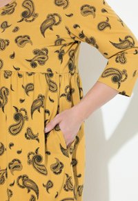 Una mano femminile nella tasca di un vestito giallo senape con stampa paisley nera e maniche a tre quarti contro un muro bianco.
