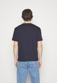 Námořnické bavlněné tričko s kulatým výstřihem a krátkými rukávy, zobrazené zezadu, v kombinaci se světle modrými džíny. Jednoduchý design, bez vzorů.