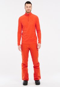Pullover in pile arancione con colletto a mezzo zip, abbinato a pantaloni da sci arancioni. Il modello indossa stivali da neve neri. Presenta una texture liscia.