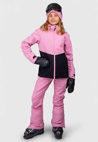 Flicka klädd i en rosa och svart skidjacka och byxor, svarta skidstövlar och vantar, rosa mössa och svarta skidglasögon på huvudet, står med handen på höften.