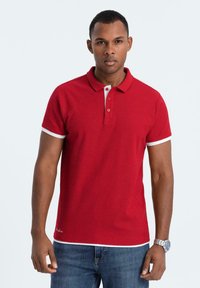 Röd polo skjorta i bomull med krage och treknappsstängning. Vita detaljer vid ärmarna och nederkanten, ihopparad med blå jeans.