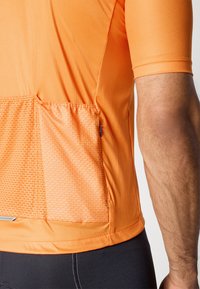 Oranžové sportovní tričko s krátkým rukávem, které má design síťkové kapsy a hladkou texturu. Obsahuje reflexní prvek ve spodní části.