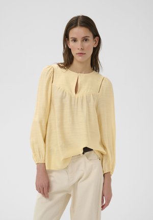 Frau trägt eine hellgelbe Bluse mit langen Ärmeln und Schlitz am Ausschnitt sowie lockere weiße Hosen, steht vor einem einfarbigen hellen Hintergrund.