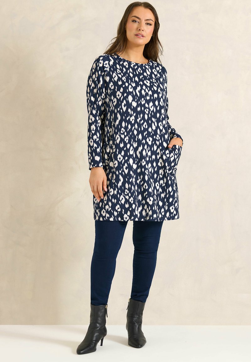 Donna che indossa una tunica a maniche lunghe con motivo blu navy e bianco, leggings blu navy e stivali neri alla caviglia, in piedi con una mano nella tasca.