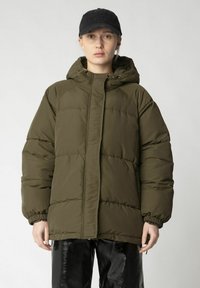 Olivgrüne Steppjacke mit Kapuze, ausgestattet mit elastischen Bündchen und einem Reißverschluss. Getragen über schwarzen, glänzenden Hosen.