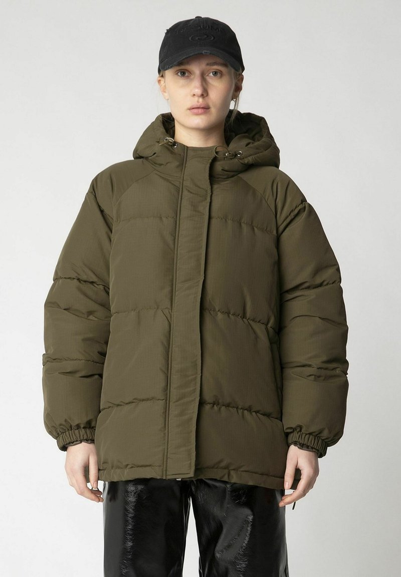 Olivgrüne Steppjacke mit Kapuze, ausgestattet mit elastischen Bündchen und einem Reißverschluss. Getragen über schwarzen, glänzenden Hosen.