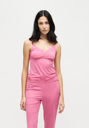 Une femme présente un caraco rose bordé de dentelle et un pantalon assorti sur un fond blanc uni.